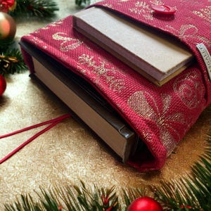 May include: A red fabric book cover with gold accents and a gold button. The cover is wrapped around a brown book with a smaller book inside.