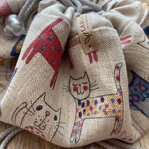 Peut inclure: Un sac à cordon beige avec un motif imprimé de chats. Le sac a une étiquette en cuir marron avec le mot "LASSIG" imprimé dessus.