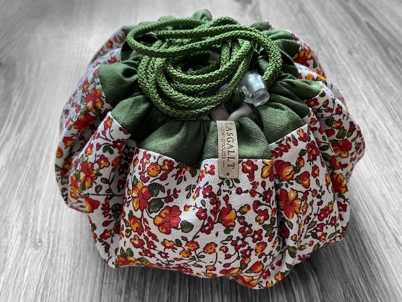 May include: A green and white drawstring bag with a floral pattern. The bag has a green drawstring and a brown tag that says "Lasgallit - linen products".