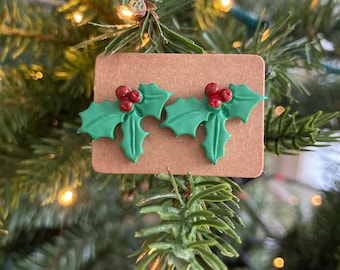 May include: A pair of green holly leaf earrings with red berry accents, displayed on a brown card. The earrings are set against a backdrop of a Christmas tree with soft, blurred lights, evoking a festive holiday theme.