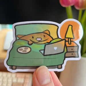 May include: A sticker featuring a cartoon animal in bed with a laptop and biscuits. The animal is brown with a white tummy and a pink nose. The bed is green and has a white blanket. There is a lamp on a bedside table next to the bed.