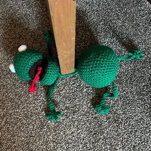 May include: A green crocheted frog toy with a red tongue and white eyes lying on a grey carpet next to a wooden post.