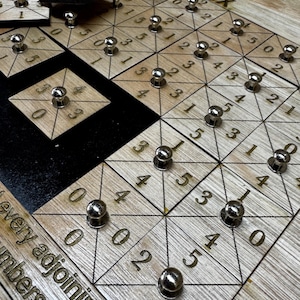 Op de afbeelding: Een houten Tetravex puzzelspel met een lichte houtnerf afwerking. Het spelbord heeft een raster van driehoeken en vierkanten met cijfers en metalen pinnen. De spelstukken zijn ook van hout met metalen pinnen.