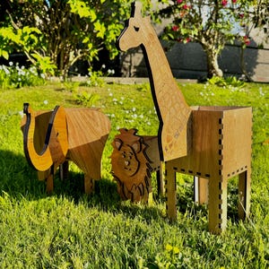 May include: Three wooden animal planters, a giraffe, elephant, and lion, stand on grass. The giraffe has a long neck, the elephant has large ears, and the lion has a mane. The planters are made of light brown wood and are suitable for a garden.