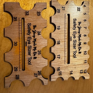 Op de afbeelding: Twee houten veiligheidsoogmaten in lichtbruin. Elk gereedschap heeft genummerde gaten en een liniaal in millimeters. De tekst "The Sheep Made Me Do It Safety Eye Sizer Tool mm" staat op elk gereedschap.