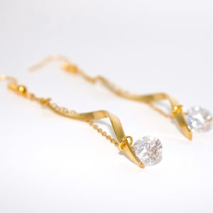May include: A pair of gold-coloured drop earrings with a swirling design. Each earring features a clear, round, sparkling gemstone at the bottom. The earrings are on a white background.