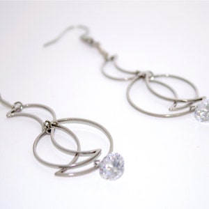 May include: A pair of silver dangle earrings with a clear crystal at the bottom of each earring. The earrings feature a crescent moon design with a circle around it.