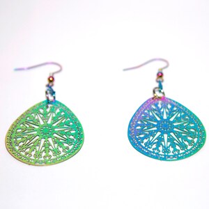May include: A pair of iridescent earrings with a teardrop-shaped design. The earrings feature a delicate, intricate pattern with a circular mandala in the centre.