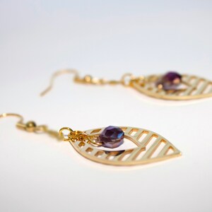 May include: Gold dangle earrings with a purple crystal bead hanging from a leaf-shaped design.