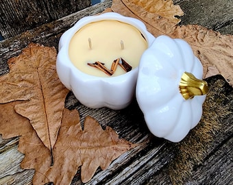 Puede incluir: Portavelas de calabaza de cerámica blanca con tapa. La vela es de color amarillo claro y tiene dos mechas. La tapa tiene un acento dorado en la parte superior. El portavelas está sobre una superficie de madera con hojas marrones.