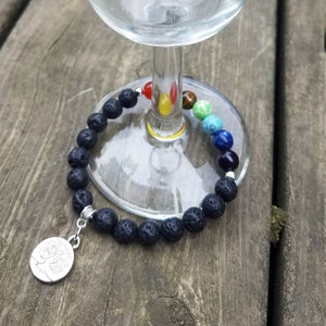 May include: A black lava rock bracelet with a silver charm featuring a tree of life design. The bracelet is wrapped around a clear glass goblet.