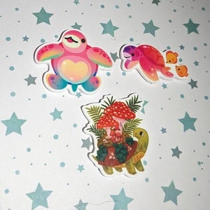 Peut inclure: Trois autocollants colorés représentant une tortue rose et bleue, une tortue rouge et orange avec deux petites tortues jaunes et une tortue brune portant un groupe de champignons rouges.