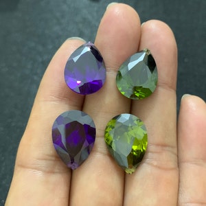 May include: Four pear-shaped gemstones, two purple and two green, are displayed on a hand. The gemstones are faceted and have a high shine.
