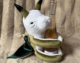 May include: A plush dragon toy with a white head, olive green body, and dark green satin wings, designed to hold a glass bottle. The bottle features a clear glass neck and a label with the text Gerard Bertrand Cote des Roses.
