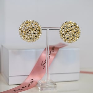 May include: A pair of gold and white round earrings with a delicate, intricate design. The earrings are displayed on a clear stand with a pink ribbon.