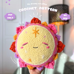 May include: A round crochet sun pillow with a pale yellow face, closed eyes, and a small smile. The sun features orange, pink, and purple rays around the edge. Text on the image reads: YUNA the dreamy sun pillow CROCHET PATTERN PDF ONLY NO SEW.