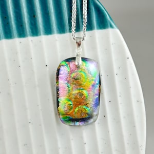 Peut inclure: Pendentif rectangulaire en verre dichroïque sur une chaîne en argent. Le pendentif présente un éventail de couleurs vives, notamment rose, jaune, vert et bleu, avec un effet irisé chatoyant. Le collier est présenté sur une assiette en céramique blanche et turquoise.