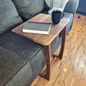 May include: A walnut wood side table with a curved design. The table has a rectangular top and is sitting on a gray couch. A book and a black coffee mug are on the table.