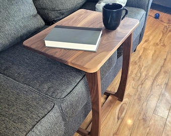 May include: A walnut wood side table with a curved design. The table has a rectangular top and is sitting on a gray couch. A book and a black coffee mug are on the table.