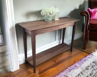May include: A dark brown wooden console table with a single shelf. The table has a simple design with straight legs and a rectangular top. The table is in a room with hardwood floors and a rug.