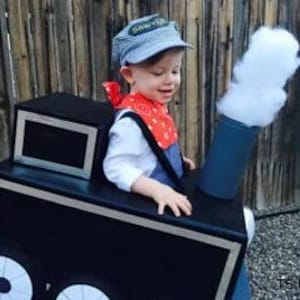 May include: A child dressed as a train conductor, wearing a blue and white striped cap and a red bandana. The child is in a black and silver cardboard train costume with a smokestack and wheels. The costume has a rectangular window.