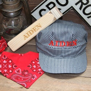 May include: A wooden whistle with the name "Aiden" engraved on it, a red bandana with a paisley pattern, and a blue and white striped engineer's cap with the name "Aiden" embroidered in red.