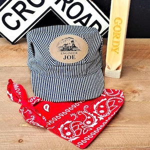 May include: A blue and white striped engineer's cap with a brown leather patch that says "Engineer Joe" and a small train image. A red bandana with a white paisley pattern is also included.