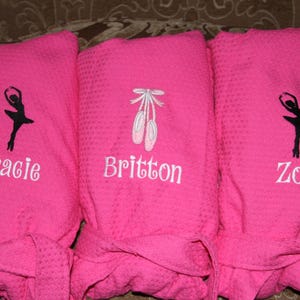 May include: Three pink bathrobes with white embroidery. The robes feature a ballerina silhouette, a pair of ballet shoes, and the names "Gracie", "Britton", and "Zoë".