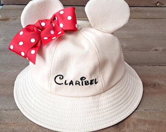 May include: A beige bucket hat with a red and white polka dot bow and the name "Claribel" embroidered on the front. The hat has two fabric mouse ears.