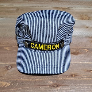 May include: A blue and white striped engineer's cap with a black leather band and the name "CAMERON" embroidered in yellow.