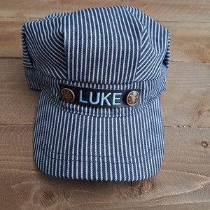 May include: A blue and white striped engineer's cap with a black leather band and a name tag that reads "LUKE".