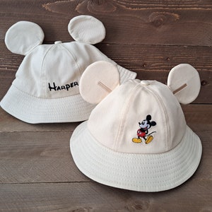 May include: Two beige bucket hats with Mickey Mouse ears. One hat has the name "Harper" embroidered on the front. The other hat has a Mickey Mouse character embroidered on the front.