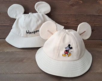 May include: Two beige bucket hats with Mickey Mouse ears. One hat has the name "Harper" embroidered on the front. The other hat has a small embroidered image of Mickey Mouse on the front.