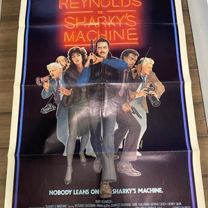 May include: A film poster for the movie "Sharky's Machine" starring Burt Reynolds. The poster features a red neon sign with the title of the film. The poster also features a group of men, including Burt Reynolds, standing in front of a dark background.
