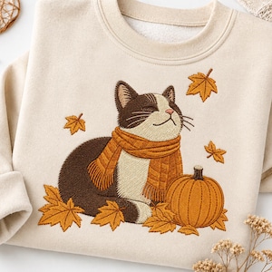 May include: A beige crewneck sweatshirt featuring an embroidered design of a brown and white cat wearing an orange scarf. The cat sits beside an orange pumpkin, surrounded by several fallen autumn leaves in shades of yellow and orange.