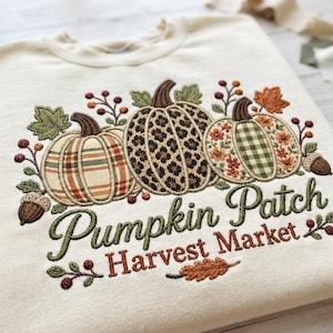 Könnte beinhalten: Ein cremefarbenes Sweatshirt mit Rundhalsausschnitt und einer Stickerei von drei Kürbissen. Die Kürbisse zeigen Karomuster, Leopardenmuster und Blumenmuster. Der Text lautet "Pumpkin Patch Harvest Market" in grünem und orangem Garn.