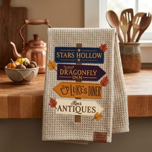 Puede incluir: Un paño de cocina beige de tejido gofrado con un gráfico de poste indicador vertical con cuatro letreros que dicen Stars Hollow, Dragonfly Inn, Luke's Diner y Kim's Antiques, decorado con hojas de otoño bordadas y una pequeña etiqueta marrón.