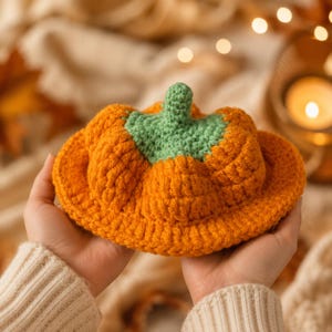 May include: A crocheted pumpkin-shaped hat in bright orange yarn with a textured, ribbed surface and a small, light green crocheted stem at the center. The hat features a wide, circular brim and a rounded crown, crafted with visible yarn stitches.