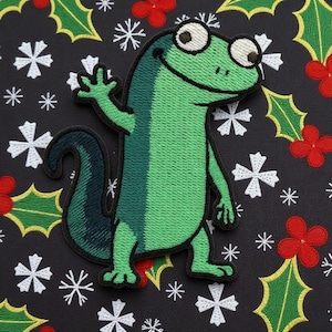 May include: A green lizard patch with a friendly expression, waving its hand. The patch is embroidered with varying shades of green and dark blue. The background is black with white snowflakes, red flowers, and green holly leaves.