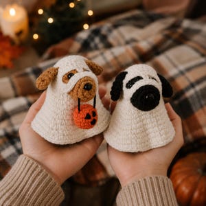 May include: Two crocheted dog figures dressed as ghosts. The left figure is a tan and white dog holding a small orange crocheted pumpkin. The right figure is a black and white dog. Both figures are wearing white crocheted ghost costumes.