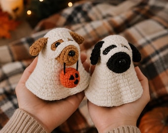 May include: Two crocheted dog figures dressed as ghosts. The left figure is a tan and white dog holding a small orange crocheted pumpkin. The right figure is a black and white dog. Both figures are wearing white crocheted ghost costumes.