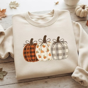 May include: A cream-colored crewneck sweatshirt featuring an embroidered design of three pumpkins. The left pumpkin has a brown and black buffalo check pattern, the center is white with small orange pumpkin prints, and the right is grey and white plaid.