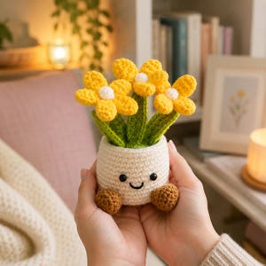 May include: A crocheted potted plant featuring three bright yellow flowers with white centres and green stems. The round, off-white pot has a stitched smiling face with black eyes and a small mouth, and two brown crocheted feet at the base.