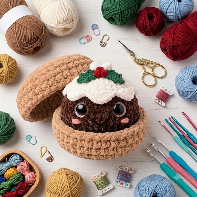 May include: A brown crocheted Christmas pudding plush with large black eyes, pink cheeks, and a white frosting top featuring green leaves and red berries. The pudding sits inside a matching brown crocheted bowl, surrounded by yarn balls and hooks.