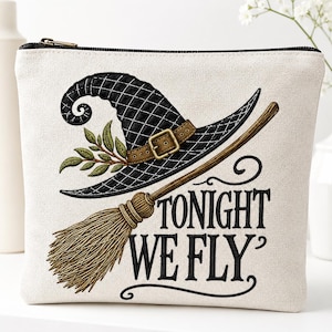 May include: A cream-colored zippered pouch featuring a black quilted witch hat with a brown buckle and green leaves, a wooden broomstick, and the black text "TONIGHT WE FLY" printed in a decorative serif font on the front of the fabric.