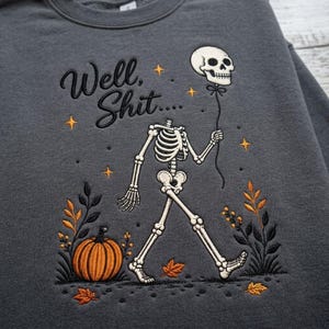 May include: A charcoal grey sweatshirt featuring an embroidered design of a walking skeleton holding a skull-shaped balloon. The text reads Well, Shit.... The graphic includes an orange pumpkin, autumn leaves, and small yellow stars.
