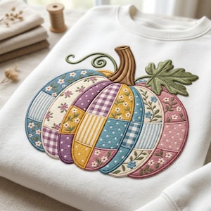 Puede incluir: Una sudadera blanca de cuello redondo con un gran diseño de calabaza bordado en el centro. La calabaza está compuesta por varios segmentos estilo patchwork en azul, amarillo, morado y rosa, decorados con motivos florales, cuadros y lunares.