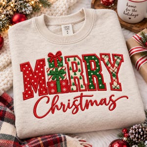 May include: A beige crewneck sweatshirt featuring embroidered text that reads Merry Christmas. The letter E is replaced by a stack of three wrapped gift boxes with bows. The letters feature red polka dots, green patterns, and plaid designs.