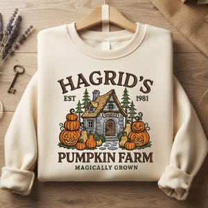 May include: A cream-colored crewneck sweatshirt featuring a graphic print of a stone cottage surrounded by pumpkins and pine trees. The text reads HAGRID'S EST 1981 PUMPKIN FARM MAGICALLY GROWN in brown lettering. The design includes carved jack-o'-lanterns.