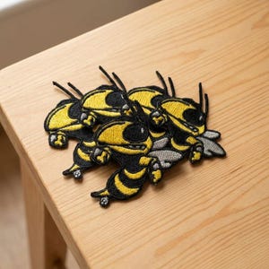 May include: A collection of three embroidered patches, each featuring a cartoon wasp with a yellow and black striped body, a large black eye, and grey wings. The patches are layered on top of each other, showcasing the detailed stitching and bold colors.